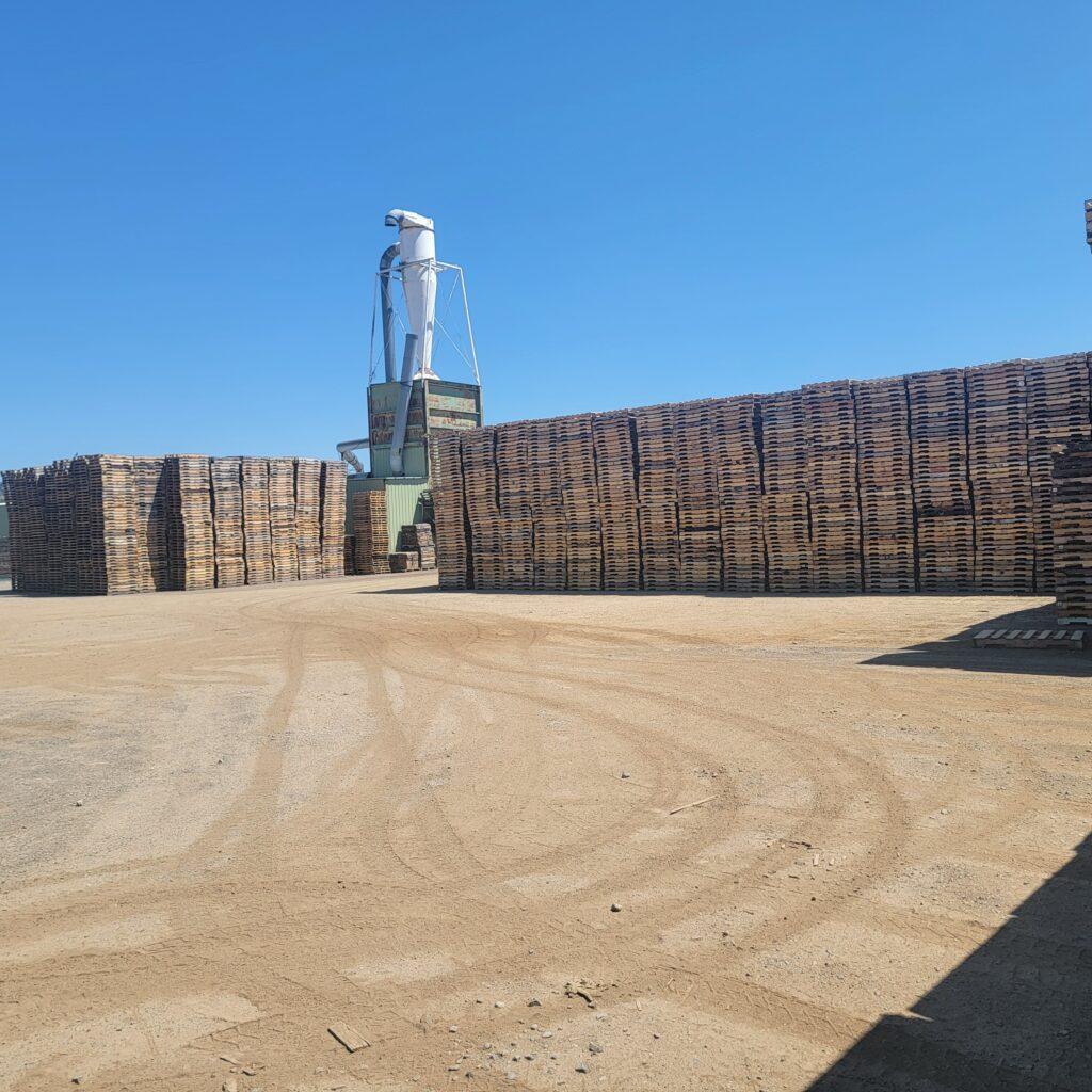 Large stacks of wooden pallets are piled high outdoors on a dirt lot under a clear blue sky. A tall industrial structure with pipes and a silo is visible in the background. Tire tracks are seen on the ground.