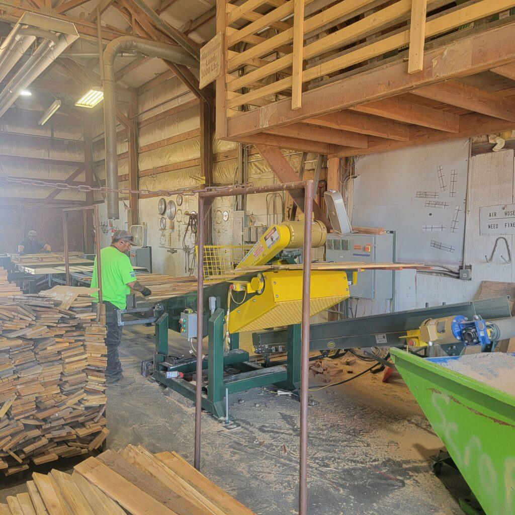 A worker in a bright green shirt operates machinery in a lumber mill. Stacks of wood are on the left, and sawdust collects below the equipment in an industrial warehouse with exposed beams and wood planks overhead.
