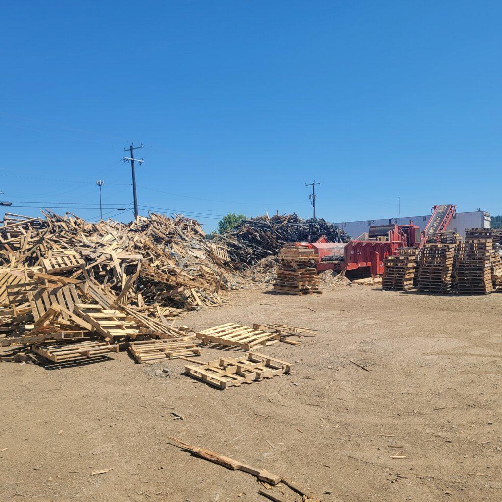 Large piles of wooden pallets and scrap wood are stacked in an open yard under a clear blue sky. Industrial machinery and buildings are visible in the background. The ground is dirt.